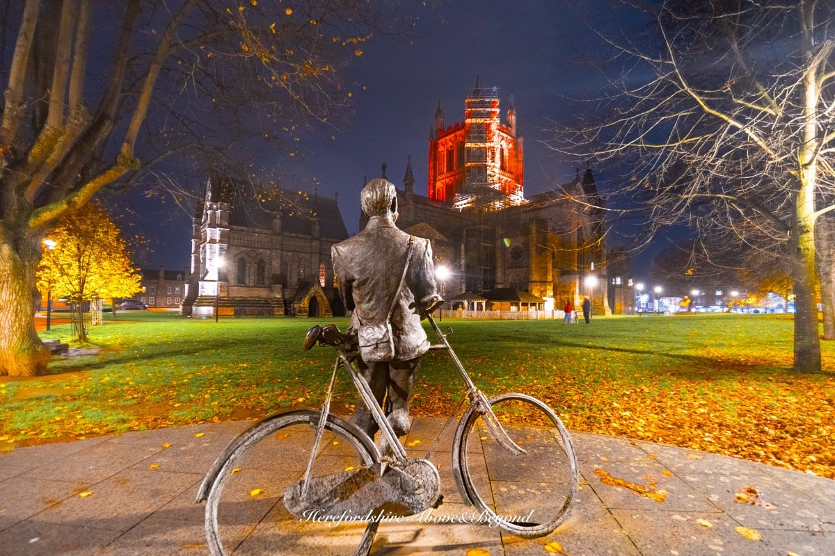 Bronze statue of a man with a bicycle looking towards Hereford Cathedral on an autumn night