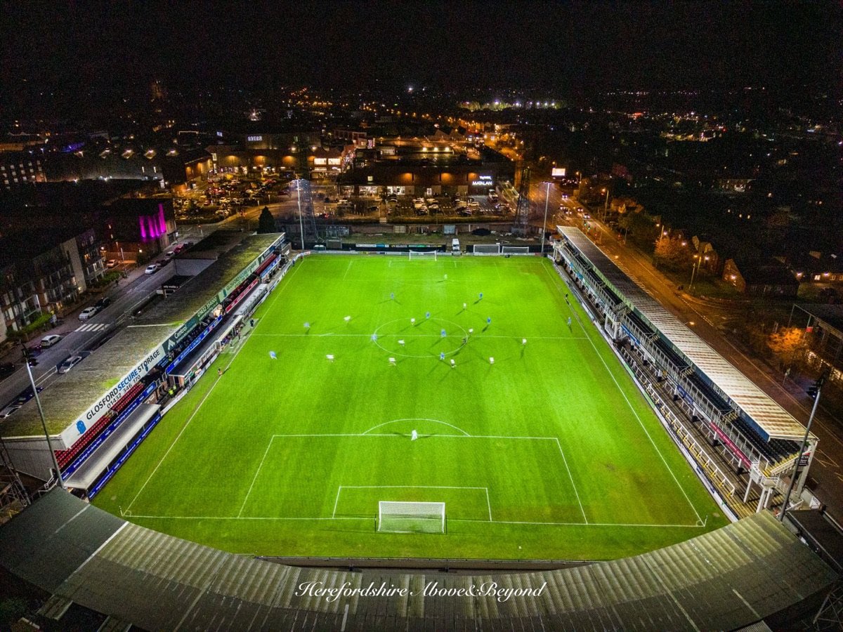 Aerial view at night of a floodlit football stadium with a match in play and the town's lights beyond